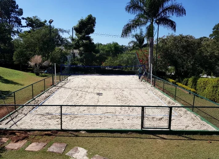 Quadra real de beach tennis com areia, rede, alambrado e vegetação ao redor.